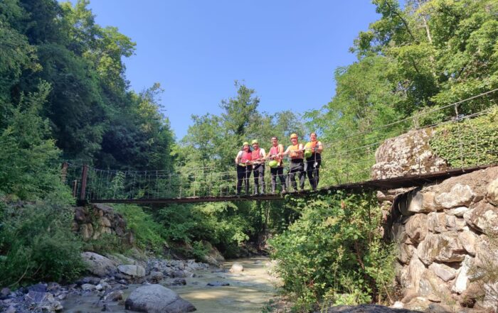 Un gruppo attrezzato con muta, caschetto e giubbotto salvagente percorre il ponte sospeso sul torrente Civasola durante l'escursione Francigena River Trekking.